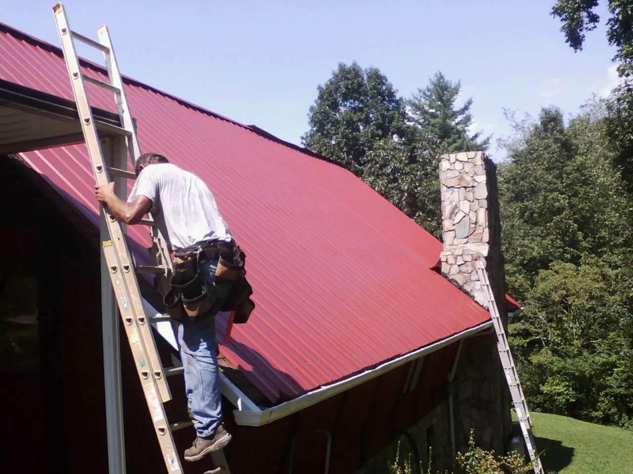 Roofer on a ladder beside a red metal roof and stone chimney — Soffit & Fascia Repair by Roofing Brothers in Mount Orab